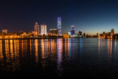 Illuminated buildings by river against clear sky at night