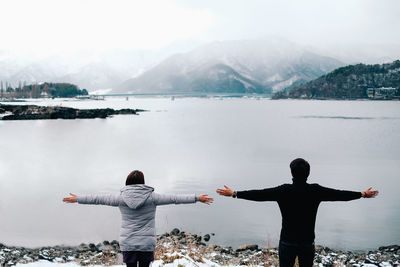 Rear view of people with arms outstretched looking at lake against mountains