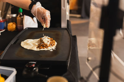 Chef sprinkles a tortilla with cheese preparing fresh quesadilla on black grill.