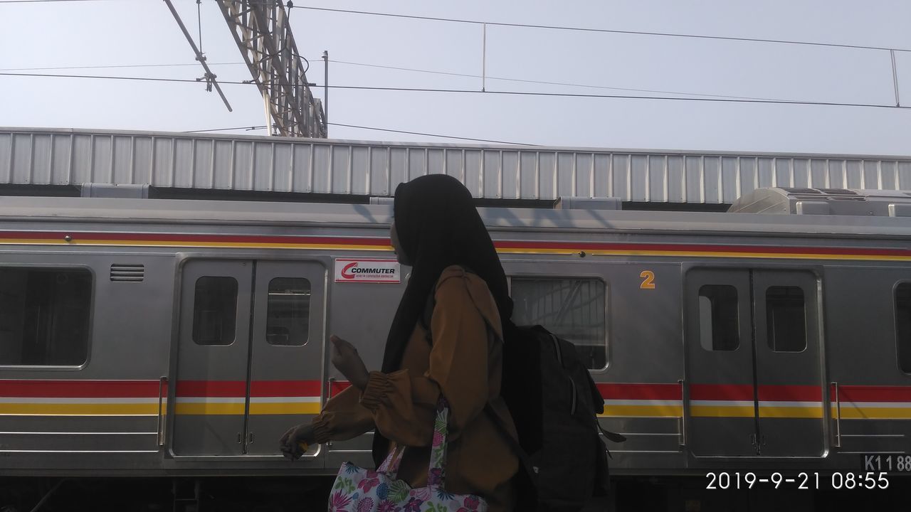 REAR VIEW OF WOMAN STANDING ON RAILROAD STATION PLATFORM