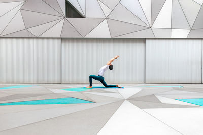 Side view of adult male in sportswear doing low lunge pose yoga exercise in spacious room with geometric wall and floor