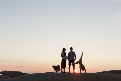 Father and mother looking at girl performing cartwheel on rock formation against sky during sunset