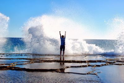 Rear view of man with arms raised standing at beach against clear sky during sunny day