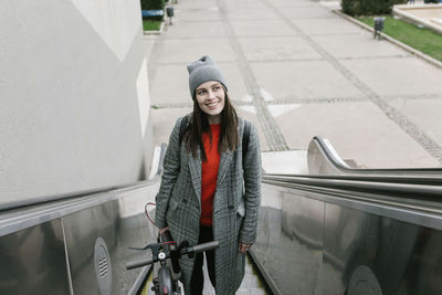 Thoughtful woman holding electric push scooter while standing on escalator in city