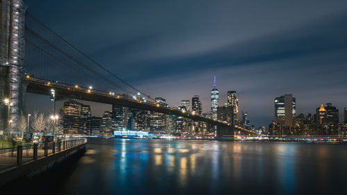 Illuminated bridge over river by buildings against sky at night