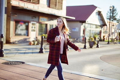 Young woman holding coffee while walking on sidewalk in city