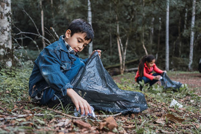 Ethnic volunteers with plastic bags picking rubbish from terrain against trees in summer woods in daylight