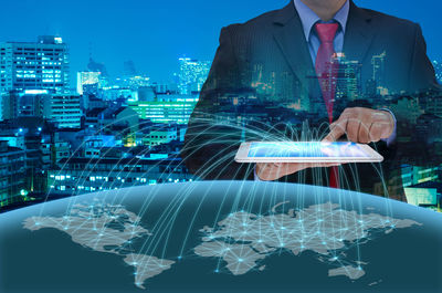 Digital composite image of businessman holding tablet showing world map against illuminated cityscape at dusk