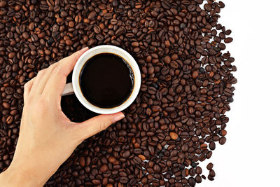 Directly above shot of hand holding black coffee on roasted beans over white background