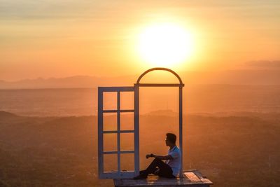 Man sitting on bench by sea against sky during sunset