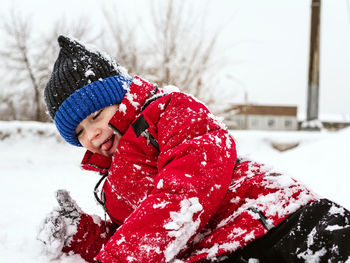Caucasian boy in a red jacket and a knitted hat is lying on the snow in the city.