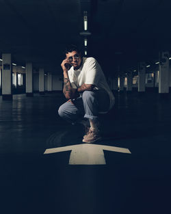 Portrait of young man wearing sunglasses crouching on arrow symbol in parking lot at night