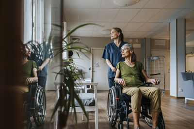 Female nurse with senior woman sitting in wheelchair and looking out of window at care home