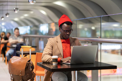 African american traveler millennial man sitting at cafe table in airport terminal, works on laptop