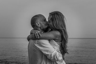 Side view of couple kissing while standing at beach against sky