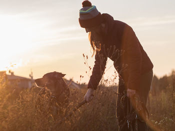 Side view of young woman with dog standing on field against sky during sunset
