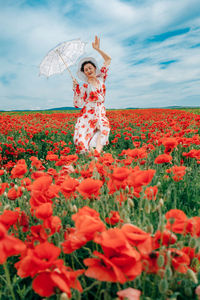 Happy woman in summer dress with a umbrella dances in the field with blooming poppies against a sky