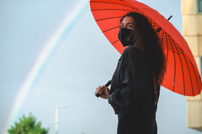 Woman with a maska and a red umbrella stands near a rainbow