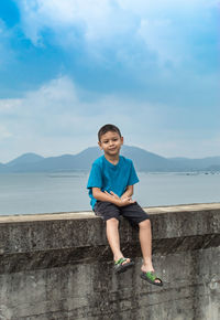 Portrait of boy sitting on retaining wall against sea 