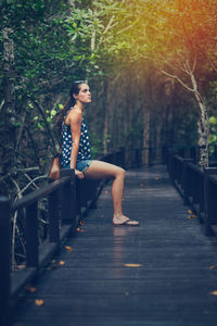 Full length of young woman sitting on footbridge against trees