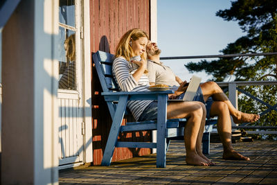 Mid adult woman using laptop while having breakfast and sitting with man at porch