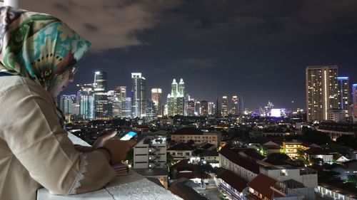 Side view of woman using phone on terrace against illuminated cityscape