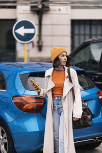 Young female with bag in stylish outfit and hat browsing on smartphone while standing on street near modern car in city