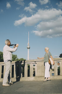 Full length of man taking photograph of senior woman while standing on bridge against tower in city