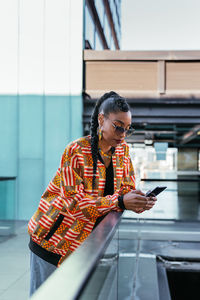 Side view of young ethnic female in stylish colorful clothes leaning on glass fence and browsing social media on smartphone on terrace outside modern building