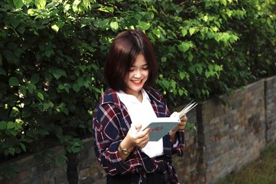 Smiling woman reading book while standing against plants