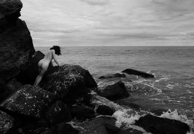 Rear view of woman sitting on rock by sea against sky