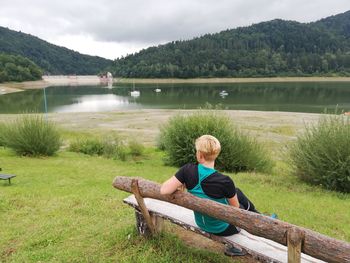 Man sitting on grass by lake against mountain