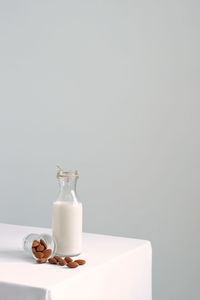 Almond milk in transparent glass bottle and pile of roast almonds on white table against white wall