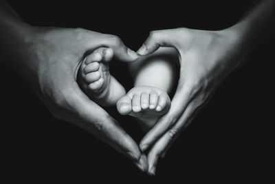 Close-up of mother hands making heart shape over baby feet against black background