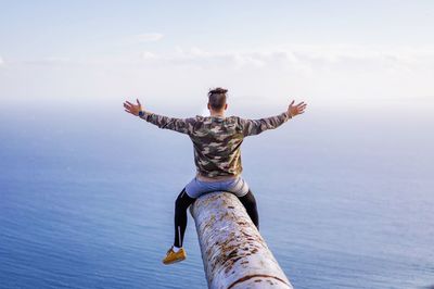 Rear view of man sitting with arms outstretched on rusty metallic pipe while looking at sea against sky