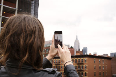 Rear view of woman photographing empire state building through smart phone in city