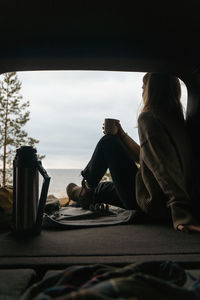 Faceless young girl in beige sweater enjoying seascape. woman sitting in car and drinking hot tea