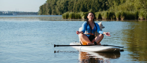 Young woman meditating on the city lake while sitting on the sup board with her dog japanese spitz