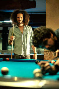 Serious ethic man with cue competing in game with player hitting billiard ball