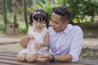 Smiling father and daughter holding piggy bank while sitting outdoors