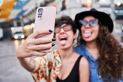 Soft focus of happy young girlfriends showing tongues and taking selfie via smartphone on weekend day on street on madrid, spain