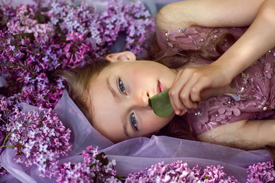 Child girl in a purple floral dress lies on the ground among lilacs on a veil in spring