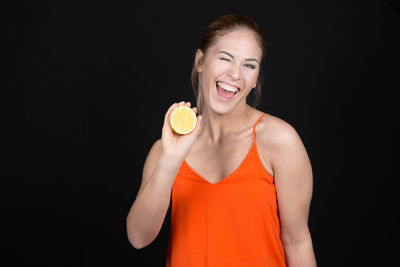 Portrait of young woman making face while holding citrus fruit against black background