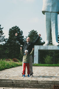 Portrait of father and son with arms raised standing at park against sky