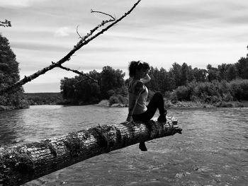 Woman sitting on tree trunk over lake against sky