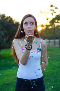 Beautiful young woman blowing confetti while standing against trees