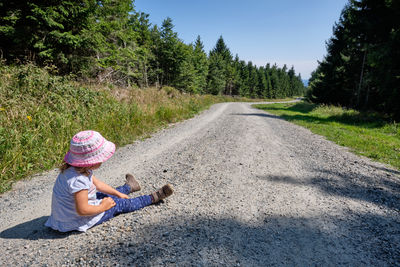 Girl sitting on road by trees against sky