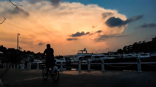 Rear view of silhouette man riding bicycle on street against sky during sunset