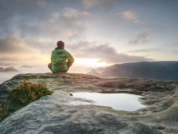 Young active woman with trekking equipment sitting, relaxing and looking at misty mountains 