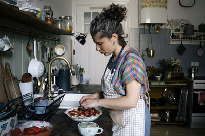 Side view of woman wearing apron garnishing pie while standing in kitchen